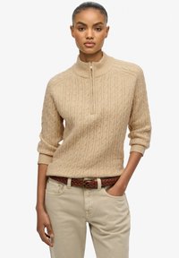 Beżowy sweter dzianinowy z wysokim kołnierzem, z suwakiem na ćwierć długości i strukturalnym wzorem w warkocz, zestawiony z jasnymi spodniami i plecionym brązowym paskiem.