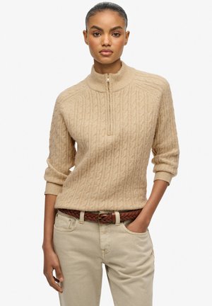 Beige gebreide trui met een hoge kraag, voorzien van een kwart rits en een gestructureerd kabelpatroon, gecombineerd met lichtgekleurde broek en een gevlochten bruine riem.