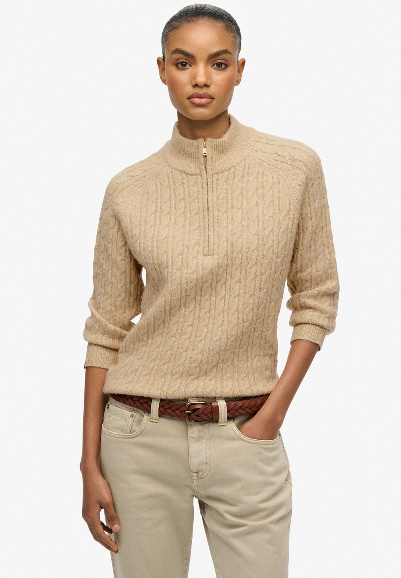 Beżowy sweter dzianinowy z wysokim kołnierzem, z suwakiem na ćwierć długości i strukturalnym wzorem w warkocz, zestawiony z jasnymi spodniami i plecionym brązowym paskiem.