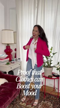 Γυναίκα που χαμογελάει και ρυθμίζει ένα φωτεινό ροζ σακάκι πάνω από λευκό πουκάμισο και τζιν σε ένα ζεστό σαλόνι με φυτά και κόκκινο βελούδινο καναπέ.