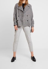 Veste double boutonnage à carreaux gris avec des revers oversize, des manches froncées et une coupe décontractée, associée à un pantalon tailleur gris clair et des mocassins noirs.