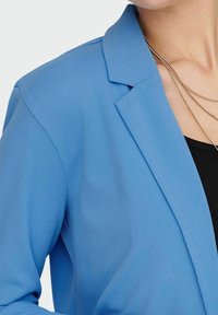 Blazer azul con cuello muesca, hecho de una tela suave. Presenta un diseño estructurado y collares de cadena en capas visibles alrededor del escote.