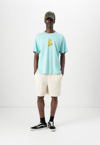 Light blue cotton t-shirt with a yellow graphic, white textured shorts, and black sneakers with white accents. Patterned cap completes the look.