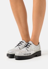 Silver glittrande oxfordskor med svarta accenter, platt gummisula, snördesign och "LOVE MOSCHINO" tryckt på sidan.