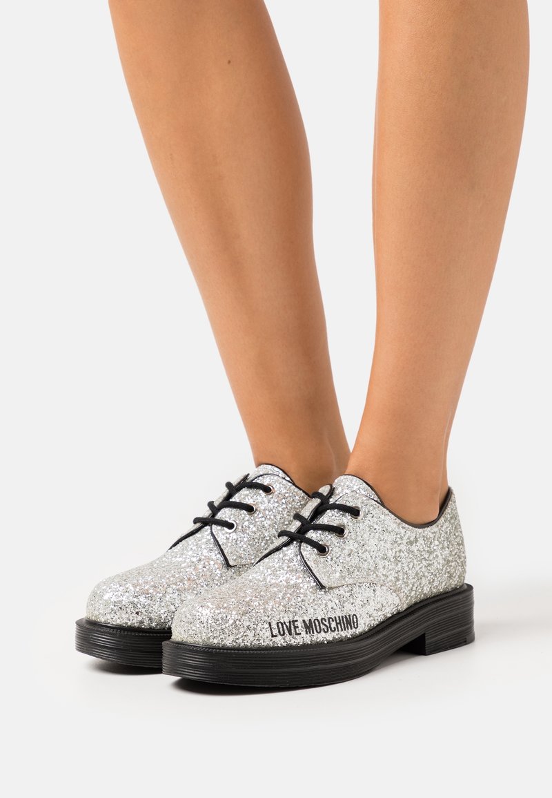 Silver glittrande oxfordskor med svarta accenter, platt gummisula, snördesign och "LOVE MOSCHINO" tryckt på sidan.