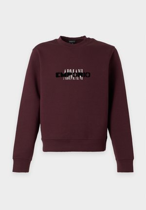 Noormees kannab maroon-värvi dressipluusi musta ja valge tekstiga, tumedad kulunud teksad ja helepruunid tossud, seisab käed taskus.