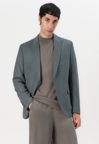 Gråtexturerad blazer med en knapp, matchad med en brun höghalsad tröja och matchande gråbyxor, vilket ger ett stilrent och modernt utseende.