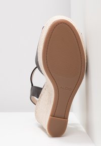 Sandale avec des brides noires et un talon compensé tressé, semelle tournée vers l’avant montrant une semelle en caoutchouc texturée et la marque "ALDO".