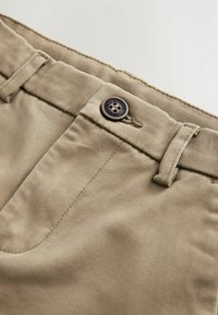 Beige bomullbyxor med slät front, med en mörk knapp märkt "AUTHENTIC" och bältesöglor med subtil textur.