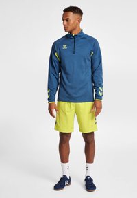Maglia blu a maniche lunghe con zip e dettagli verde lime, abbinata a pantaloni corti verde lime. Presenta un tessuto texturizzato e un design sportivo.