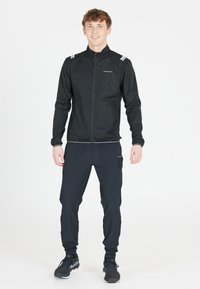 Zwarte sportjack met ritssluiting aan de voorkant, lange mouwen, reflecterende schouderaccenten, gecombineerd met zwarte taps toelopende broek en sportschoenen.