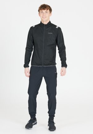 Veste légère noire avec col haut et fermeture éclair, dotée d'accents réfléchissants. Associée à un pantalon noir ajusté et des chaussures de course.