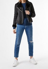 Giacca nera in similpelle scamosciata con colletto in pelliccia, indossata sopra un maglione blu a maglia e abbinata a jeans blu strappati e sneakers bianche.