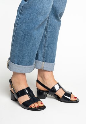 Black patent leather heeled sandals with open toe, featuring geometric cutouts, adjustable strap, and block heel, paired with cuffed denim jeans.
