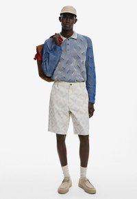Blau-weißer gestreifter Langarmshirt, creamfarbene gemusterte Shorts, beige Schuhe und eine beige Kappe, mit einem braunen Rucksack mit roten Akzenten.