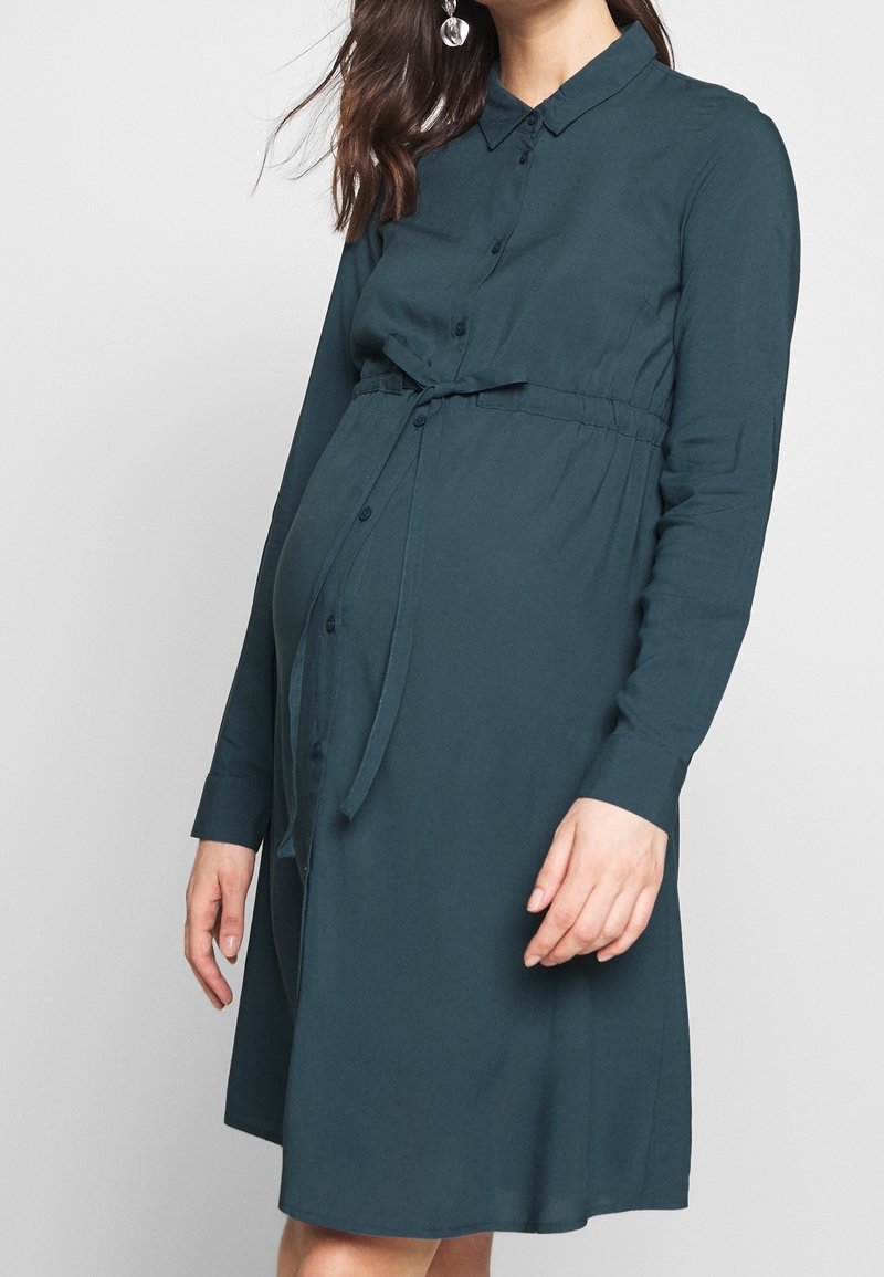 Femme portant une robe de maternité bleu sarcelle foncé à manches longues avec des boutons et une ceinture à la taille, debout sur un fond clair uni.