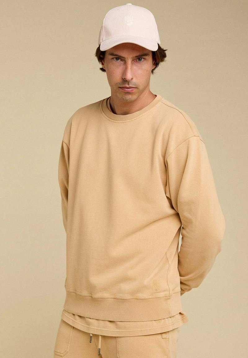 Sweatshirt beige avec un col rond, des coutures d'épaule tombantes et des poignets côtelés. Associé à une casquette rose pâle arborant un logo.