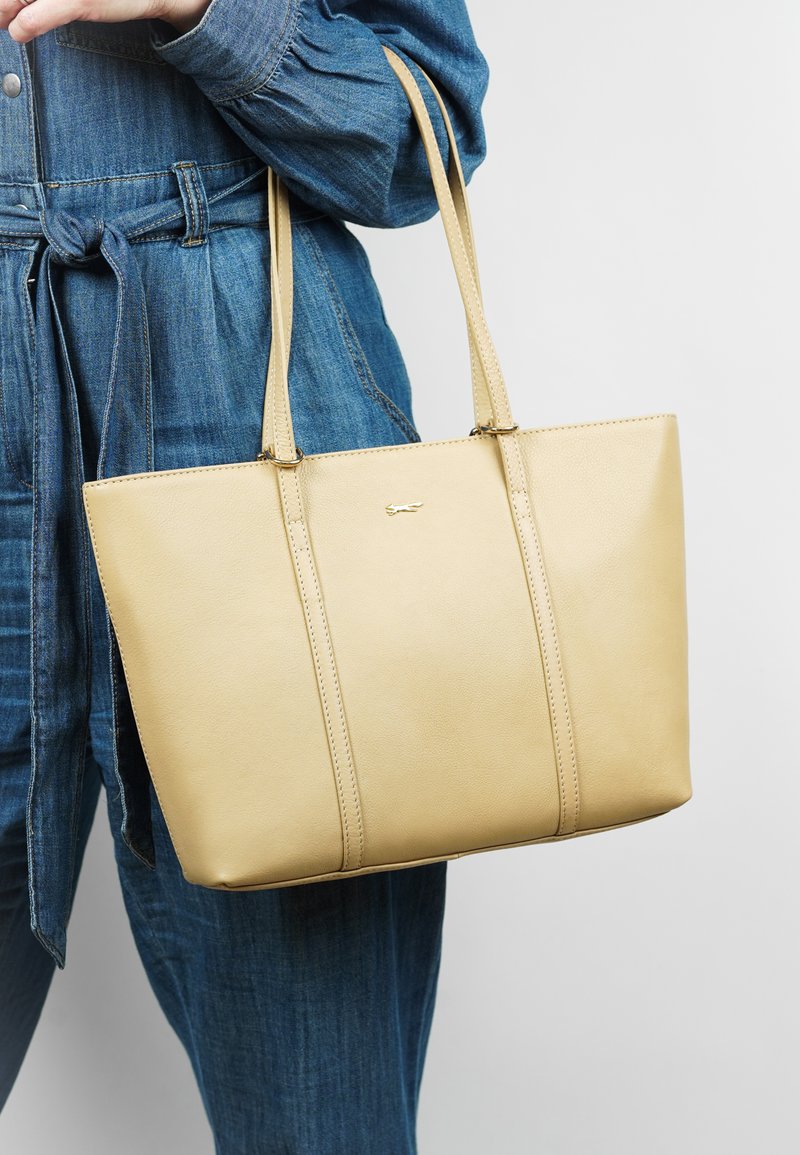Sac fourre-tout en cuir beige avec deux poignées supérieures, présentant une texture lisse et des détails de couture subtils. Design simple et forme structurée.