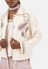 Chaqueta bordada de color beige claro con patrones florales en diseño paisley, cierre de botones y acentos a rayas. Lleva una blusa clara debajo.