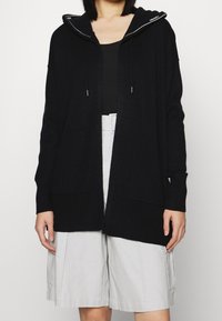 Cardigan in maglia nera con cappuccio, tasche frontali e cordini. Abbinato a pantaloni corti grigi chiari. La texture appare morbida e accogliente.