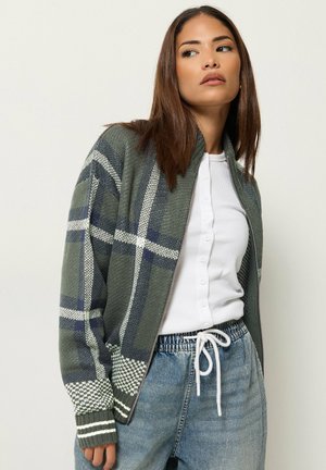 Femme portant un cardigan à carreaux verts zippé, une chemise blanche boutonnée et un pantalon en jean bleu avec cordon, regardant vers sa gauche sur un fond uni.
