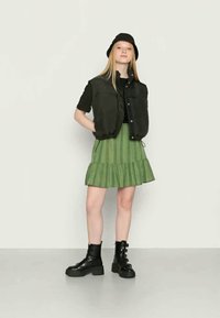 Jovem mulher com um chapéu preto estilo bucket, colete acolchoado sem mangas preto, blusa preta, saia verde com padrão e botas de combate pretas, de pé com as mãos atrás das costas.