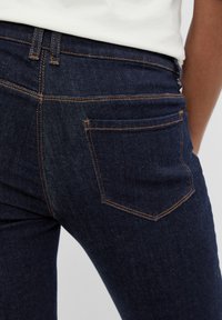 Vaqueros de denim azul oscuro con diseño ajustado, que presentan costuras naranjas y un bolsillo trasero con forma redondeada. Textura suave.