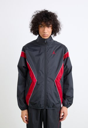 Veste coupe-vent noire avec des accents rouges et un passepoil blanc, dotée d'un col haut et d'un logo sur la poitrine, fabriquée à partir d'un matériau léger.