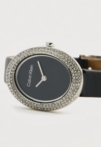 Αυτή είναι μια οβάλ μαύρη ρολόι που διαθέτει ασημένιο bezel με κρύσταλλα, μινιμαλιστικούς λευκούς δείκτες και μαύρο δερμάτινο λουράκι.