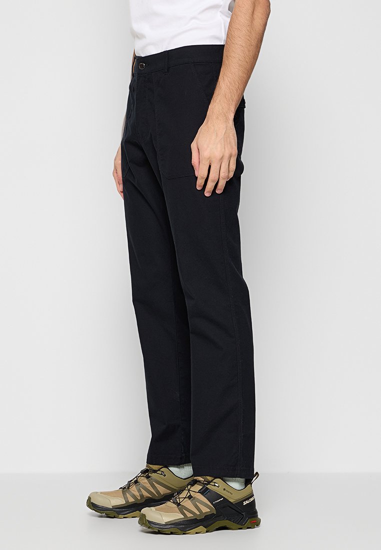 Mann in schwarzen, geraden Hosen, weißen Socken und tanfarbenen sowie schwarzen Wanderschuhen, der vor einem einfarbigen hellgrauen Hintergrund steht.