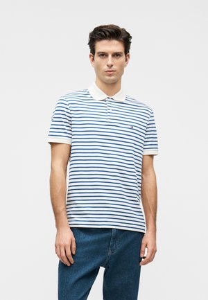Giovane uomo che indossa una polo bianca con righe orizzontali blu e jeans blu, in piedi davanti a uno sfondo chiaro e uniforme.