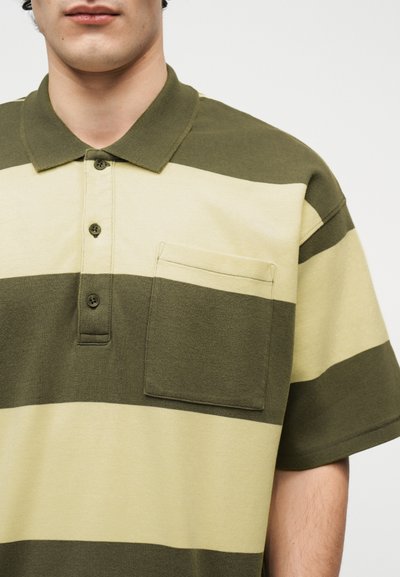 Άνδρας που φοράει κοντομάνικο πόλο με πράσινες και μπεζ φαρδιές ρίγες, με τρία κουμπιά και τσέπη στο στήθος, σε λευκό φόντο.