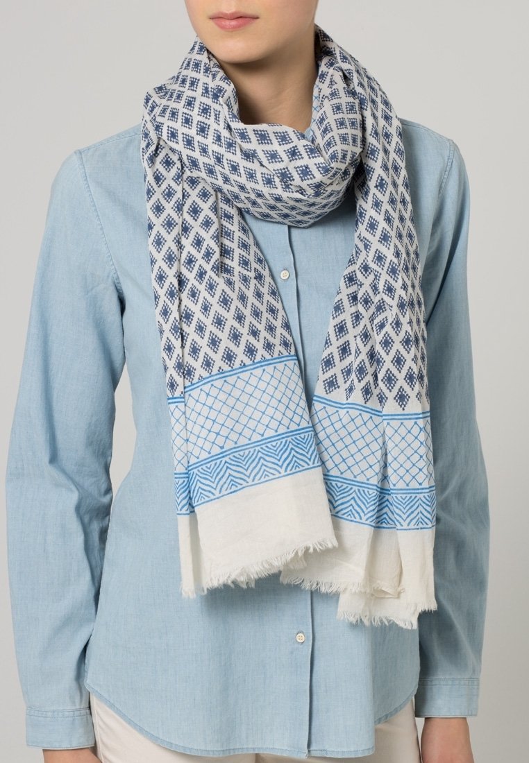 Femme portant une écharpe bleue à motifs sur une chemise bleu clair à boutons et un pantalon beige sur un fond neutre.