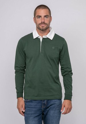 Homme aux cheveux courts et barbe portant un polo à manches longues vert foncé avec un col blanc et un jean bleu, debout devant un fond uni.