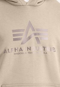 Sudadera beige con una textura suave, que presenta un logo reflectante y texto en gris que dice "ALPHA INDUSTRIES, KNOXVILLE, TENNESSEE, U.S.A."