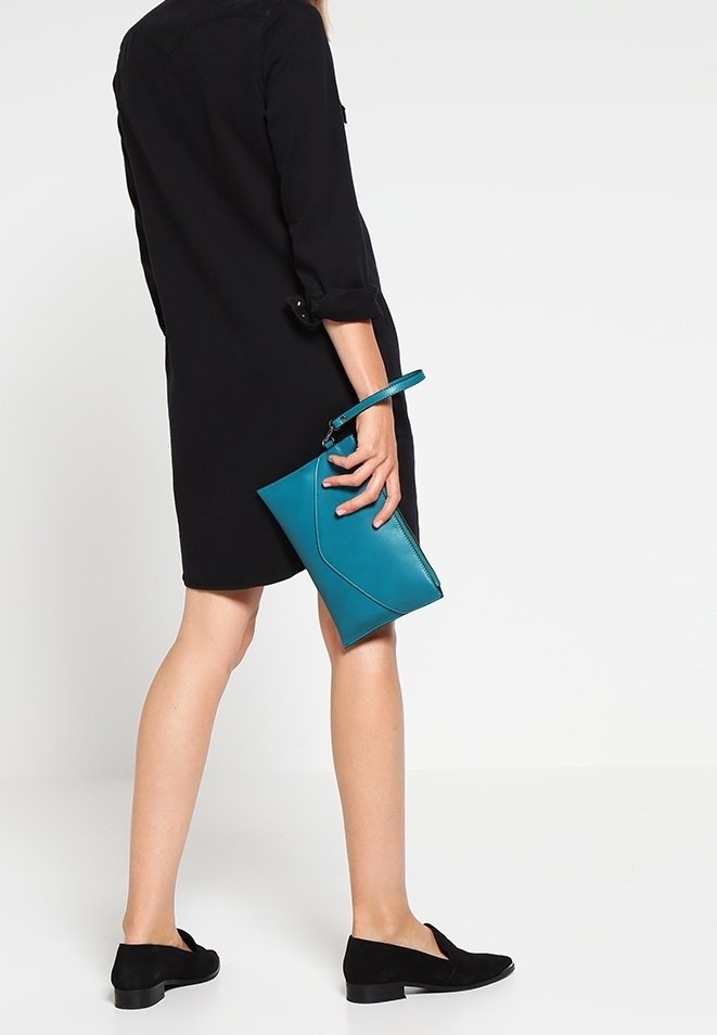 Femme en robe noire et ballerines noires tenant une pochette bracelet bleu sarcelle sur un fond blanc uni.