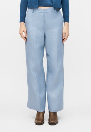 Γυναίκα που φοράει ανοιχτό μπλε παντελόνια με φαρδιά πόδια, μπλε κοντό πουλόβερ και καφέ μποτάκια αστραγάλου, στέκεται μπροστά σε λευκό φόντο.