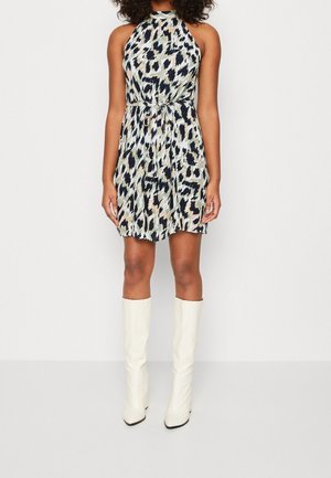 Donna che indossa un vestito senza maniche lungo fino al ginocchio con stampa animalier e vita stretta, stivali bianchi al ginocchio, in piedi su sfondo uniforme.