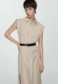 Abito senza maniche beige con colletto, fessure laterali e cintura nera a contrasto. Tessuto liscio, vestibilità su misura, design minimale.