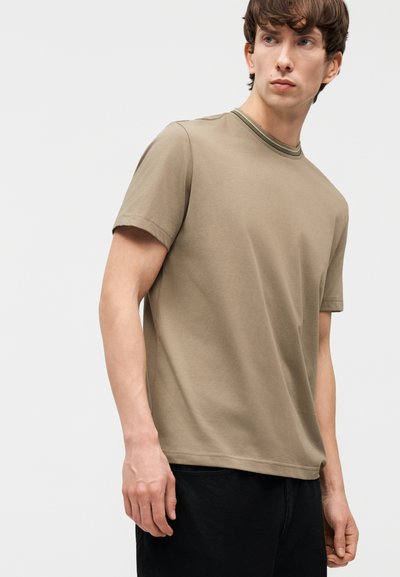 Νέος άνδρας με μπεζ κοντομάνικη μπλούζα με ριγέ λαιμόκοψη και μαύρο παντελόνι, κοιτάζει στο πλάι με φόντο μια απλή επιφάνεια.