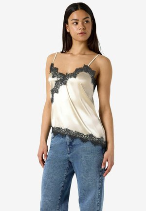 Femeie purtând un maiou bej din satin cu margine din dantelă neagră și blugi albaștri cu talie înaltă, stând cu mâinile relaxate pe lângă corp.