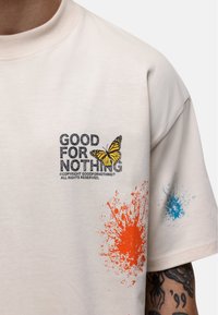 Kremowa koszulka bawełniana z graficznym nadrukiem z napisem "DOBRZE NA NIC," żółtą motylkiem oraz pomarańczowymi i niebieskimi akcentami w postaci splat.