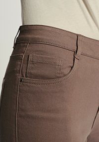 Brązowe dżinsy o dopasowanym kroju, z małą tylną kieszonką i detalami stebnów. Gładka struktura i prosty design.