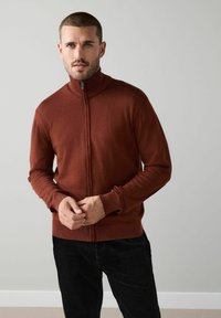 Barna cipzáros pulóver magas gallérral, puha anyagból. Hosszú ujjakkal és bordázott mandzsettával rendelkezik. Fekete kordbársony nadrággal párosítva.