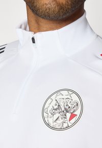 Hvid sportslig zip-up top med høj krave, sorte striber på skuldrene og et fremtrædende rundt Ajax-emblem på brystet.