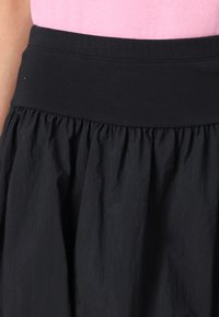 Jupe noire avec une large taille élastique et une texture froncée. Le tissu semble léger et légèrement froissé, mettant en avant le mouvement.