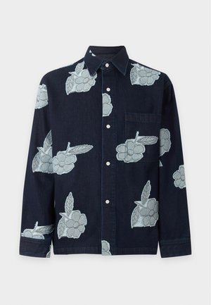 Man med glasögon, vit underställströja, marinblå skjorta med knappar och ljusblå blommönster, svarta byxor och vita sneakers som står mot en enfärgad bakgrund.