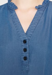 Chemise en chambray bleu avec un décolleté en V, un col froncé et quatre boutons noirs sur le devant. Texture douce, design décontracté.