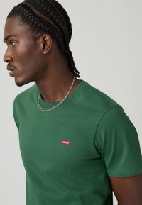 Tricou verde Levi's cu guler rotund, având un mic logo roșu pe piept, purtat cu o brățară din argint.
