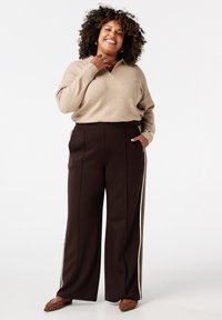 Beige trui met een v-hals, gecombineerd met bruine broek met wijde pijpen en zijstrepen. Gecombineerd met ringen en platte schoenen met luipaardprint.